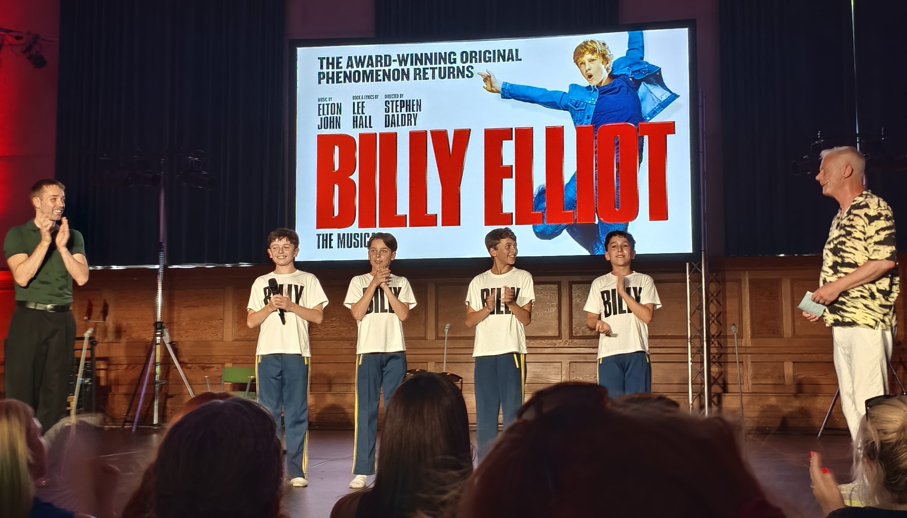 Two men stand on a stage with four young boys, all wearing white t-shirts that say 'Billy'. Above them is a screen featuring a promotional poster for Billy Elliot: The Musical