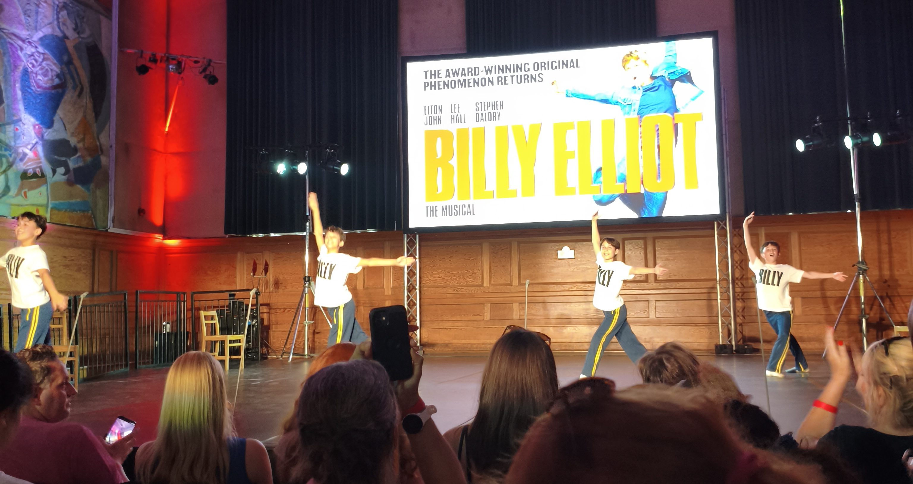 Four boys wearing white t-shirts with the word 'Billy' on them hold a ballet pose on a stage. Above them is a promotional poster for Billy Elliot: The Musical